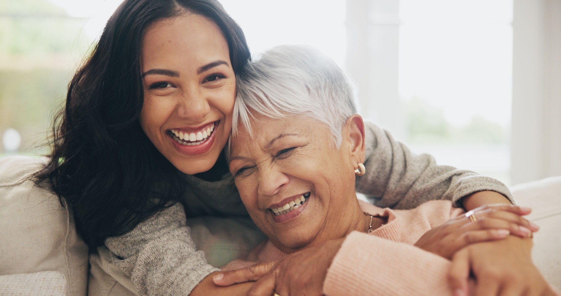 Abrazo, retrato y sonrisa de anciana con hija en el sofá en la sala de estar de la casa para vincularse o visitar. Cara, familia o amor con una persona feliz y una madre mayor abrazándose en el apartamento juntos