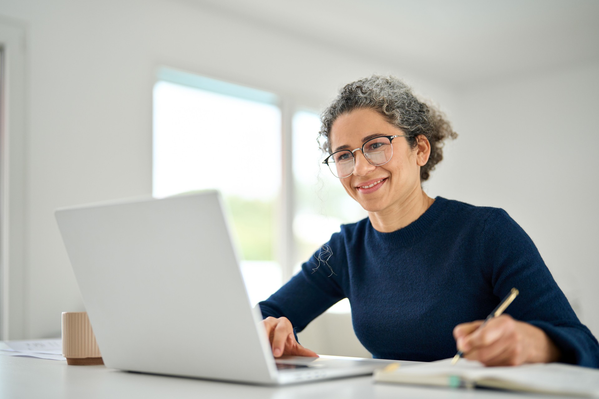 Mujer feliz de mediana edad sentada en la mesa y usando notas de escritura de computadora portátil.